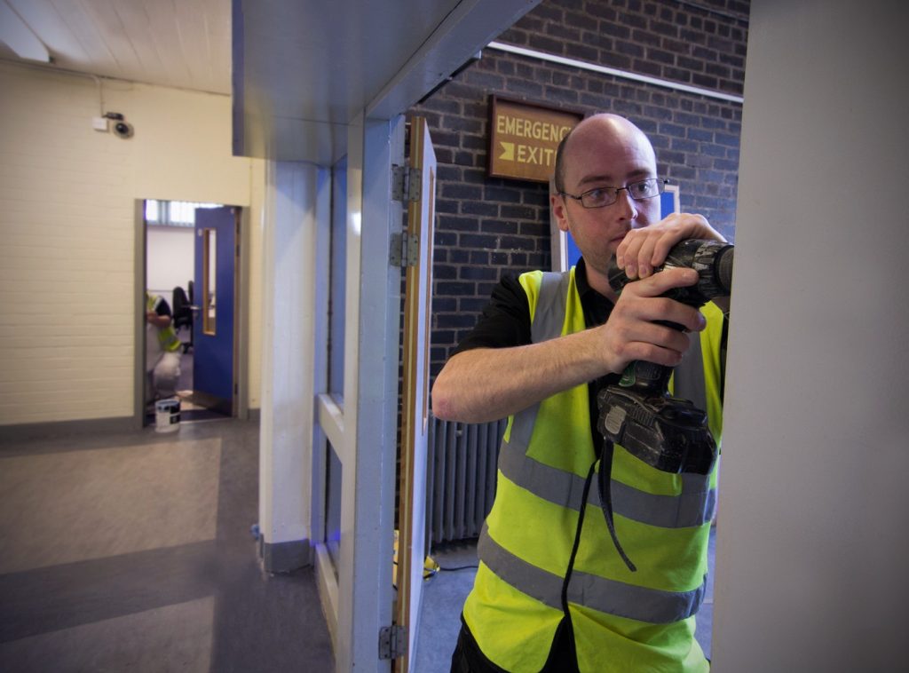 A Harvest Contracts joiner in a hi-vis vest using a cordless drill on a door frame inside a commercial building