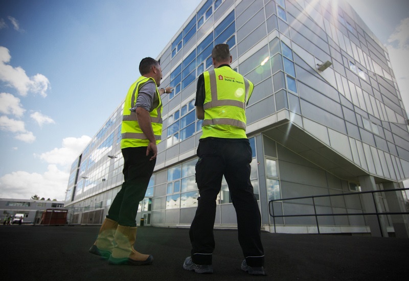 Two Harvest Contracts operatives in branded hi-vis vests looking up at a modern glazed commercial building